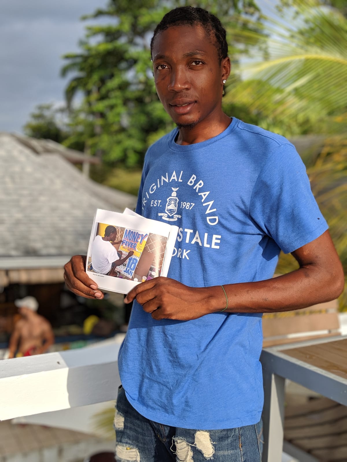 Man in a blue t-shirt holding a small publication open to a page that has a photo of him painting the 'Money Fever' sign.