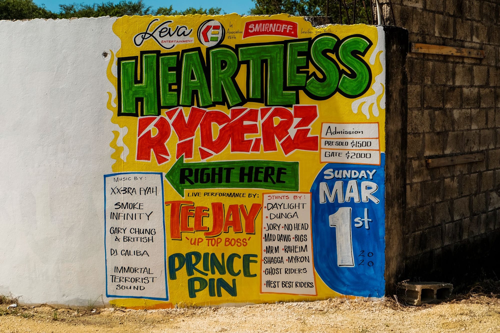 Mural sign filled with text, including "Heartless Ryderz, Right Here, Sunday, Mar 1st. Tee Jay 'Up Top Boss', Prince Pin". Other artists are also listed and there are sponsorship logos from different brands including Smirnoff.