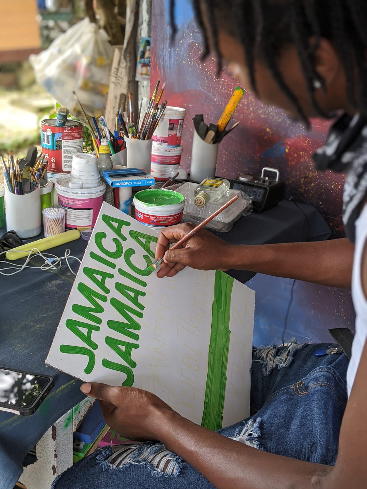 View from over the shoulder of a man painting casual green lettering onto a simple white board. Behind him a variety of painting tools and materials are visible on a table.
