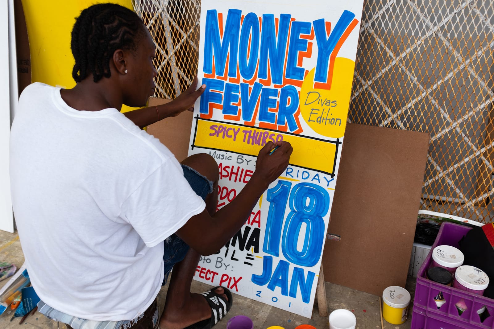 Man sat on a stool painting a sign with paint pots around him on the floor.