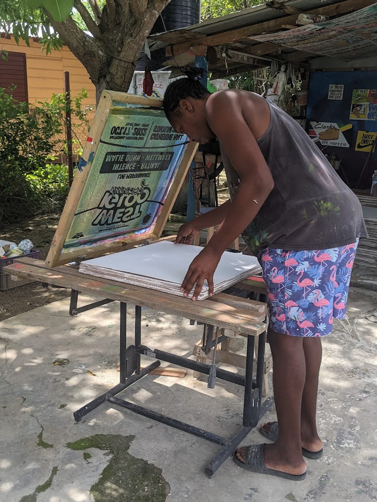 Man stooping over a screenprinting station setting the substrate in place while the hand-lettered artwork is visible in reverse on the screen that is elevated above it.