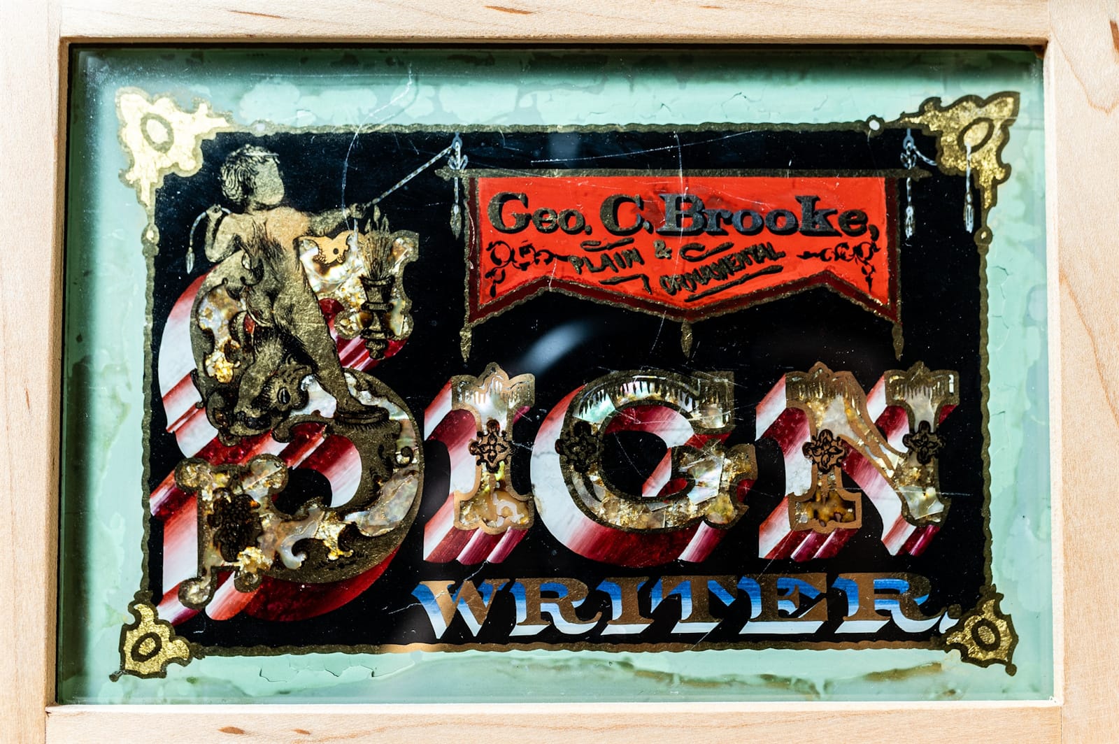 Highly decorated gilded and painted glass panel with the main text saying "Sign Writer" with a cherub depicted over the upper portion of the S. The cherub is holding a banner above the main lettering that reads "Geo. C. Brooke, Plain & Ornamental".