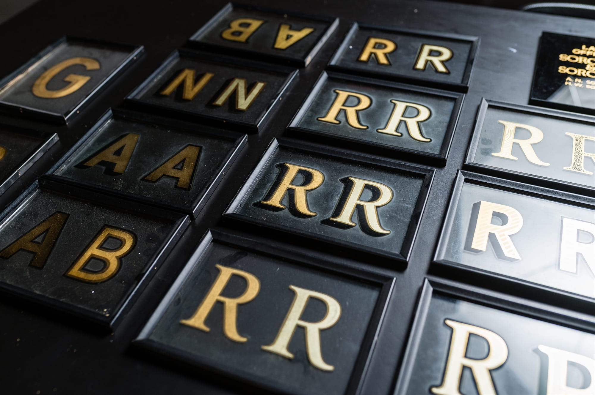 An array of framed glass panels, each with gilded letters in the centre. They are set out in an array on a black tabletop.