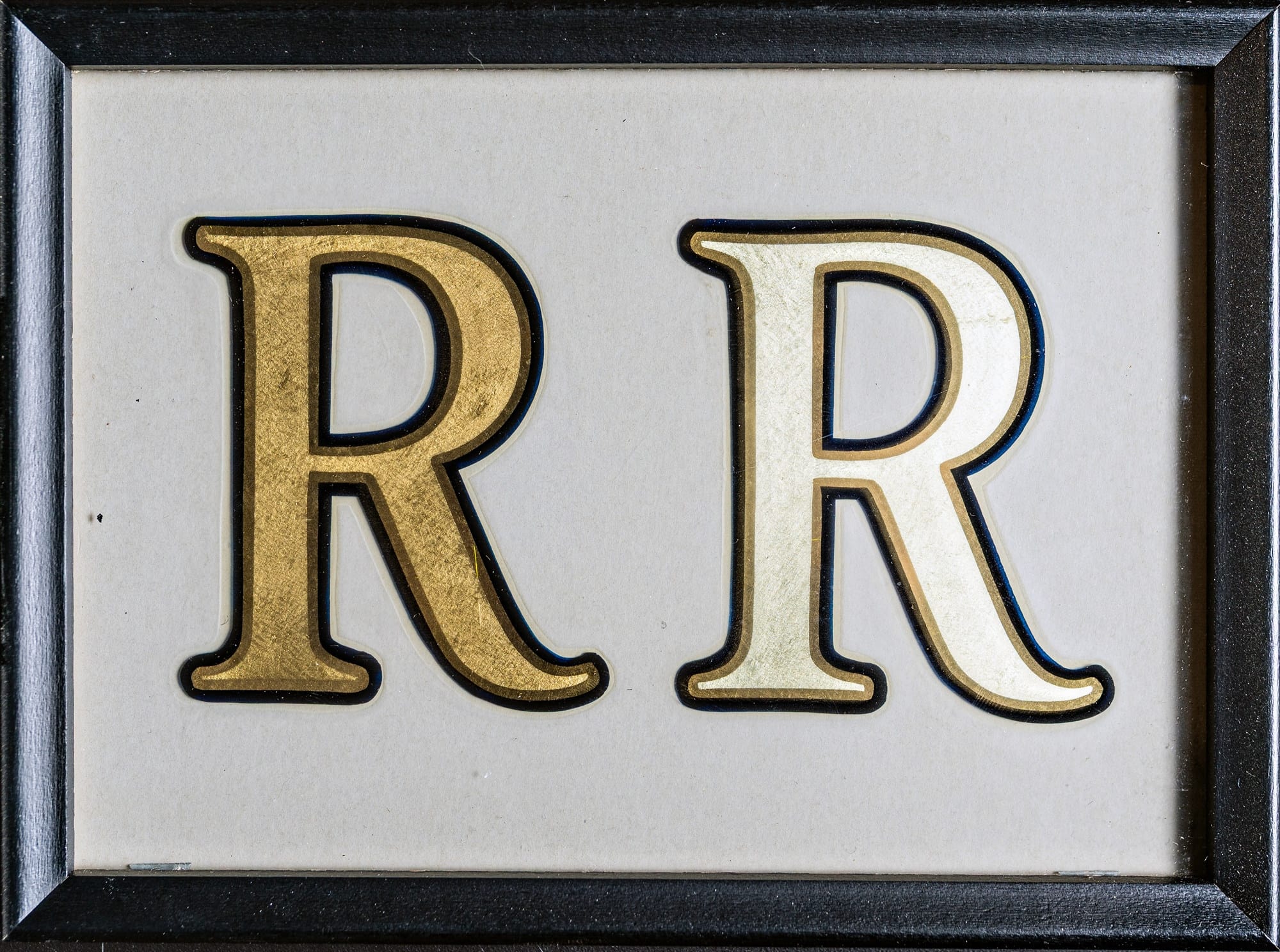 Two Roman R's with softly rounded serifs, each with a matte gold leaf effect in the centre, outlined with a shiny gild and black line. The one on the left has a darker tone of gold to the one on the right which is almost white in appearance.