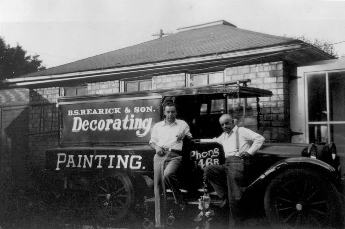 Black and white photo of two men posing in front of a vintage black truck. The truck's side panel is lettered in white with "B.S. Rearick & Son, Decorating, Painting". A phone number is painted onto the passenger door.