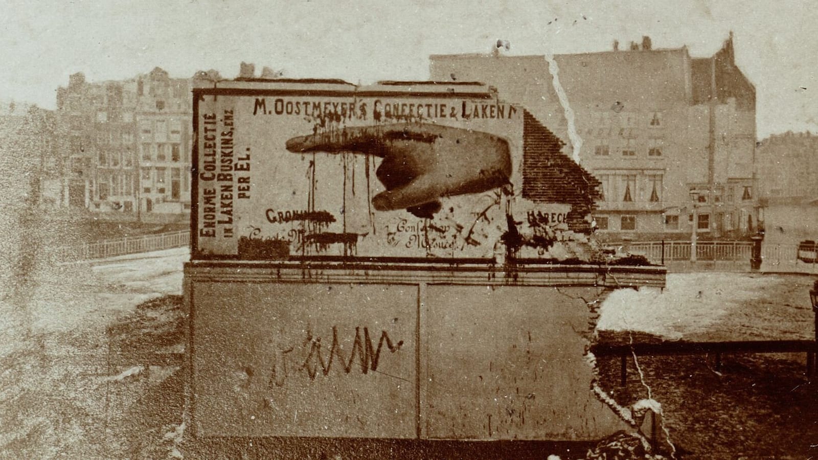 Surviving portion of a wall with derelict land behind and then buildings behind that. The wall has a huge pointing hand painted on it and the name of the business (M. Oostmeyer's Confectie & Laken...) in small letters above that.
