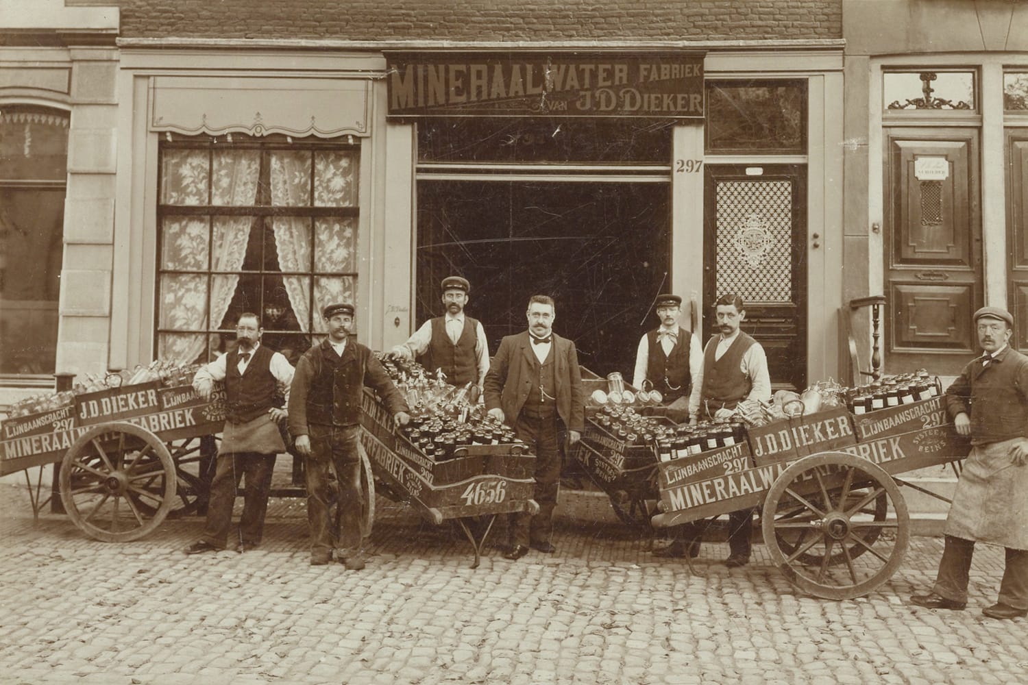 Men posing with loaded carts in front of a shopfront. The carts and the shop's fascia sign advertise "J.D. Dieker, Mineraalwater Fabriek".