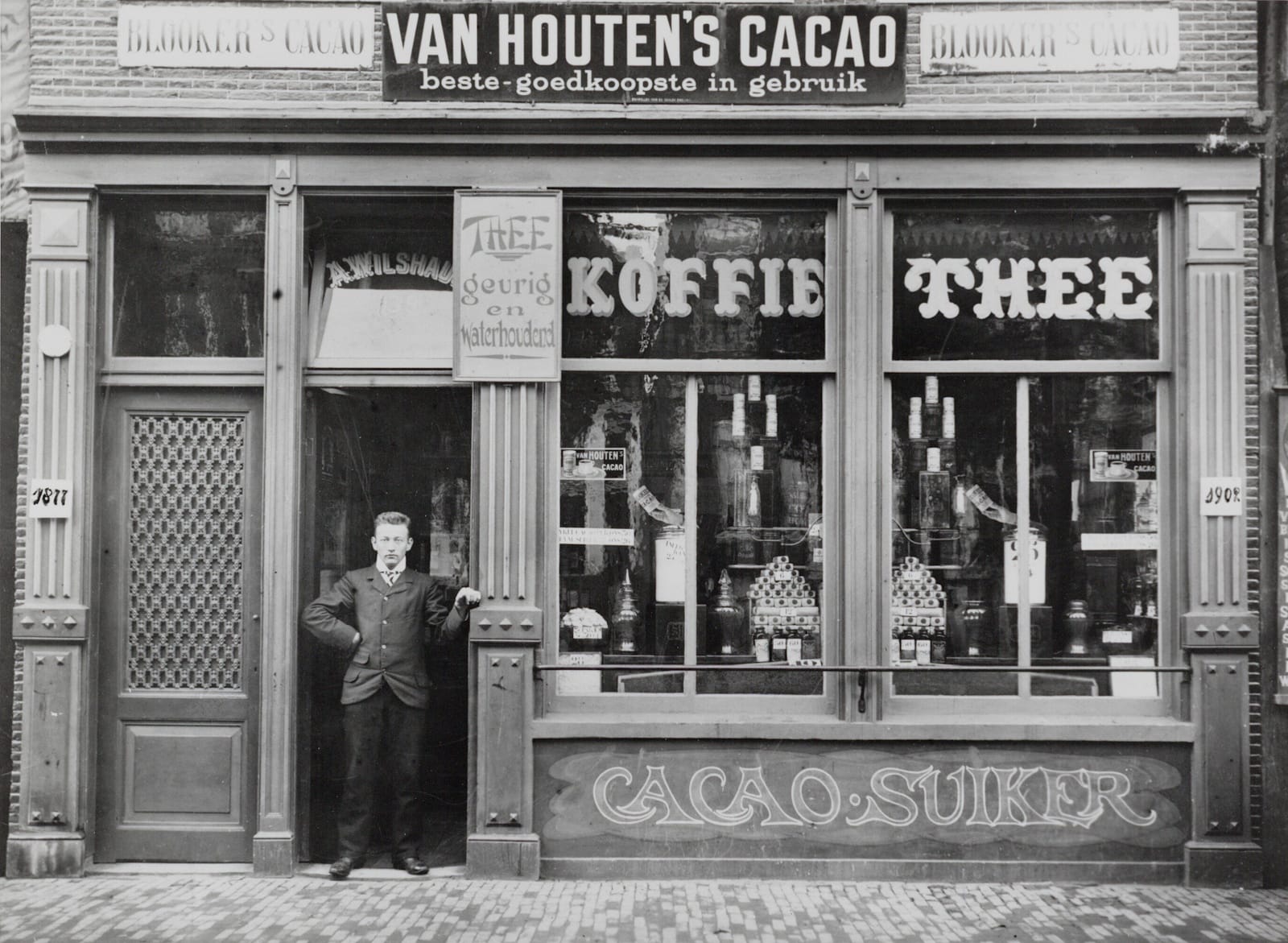 Full shopfront in a black and white photo with a man posing in the doorway. The shop has a "Cacao Suiker" sign painted along a panel beneath the windows that dominate the frontage. Above the window displays there are two transoms with "Koffie" and "Thee" painted in them respectively. There is then the business name (A.Wilshaus) painted in the transom above the door and, mounted next to this on the frame, a small tea advertisement. In the position of the fascia signs are what look like enamel signs for two cocoa brands: Van Houten's and Blooker.