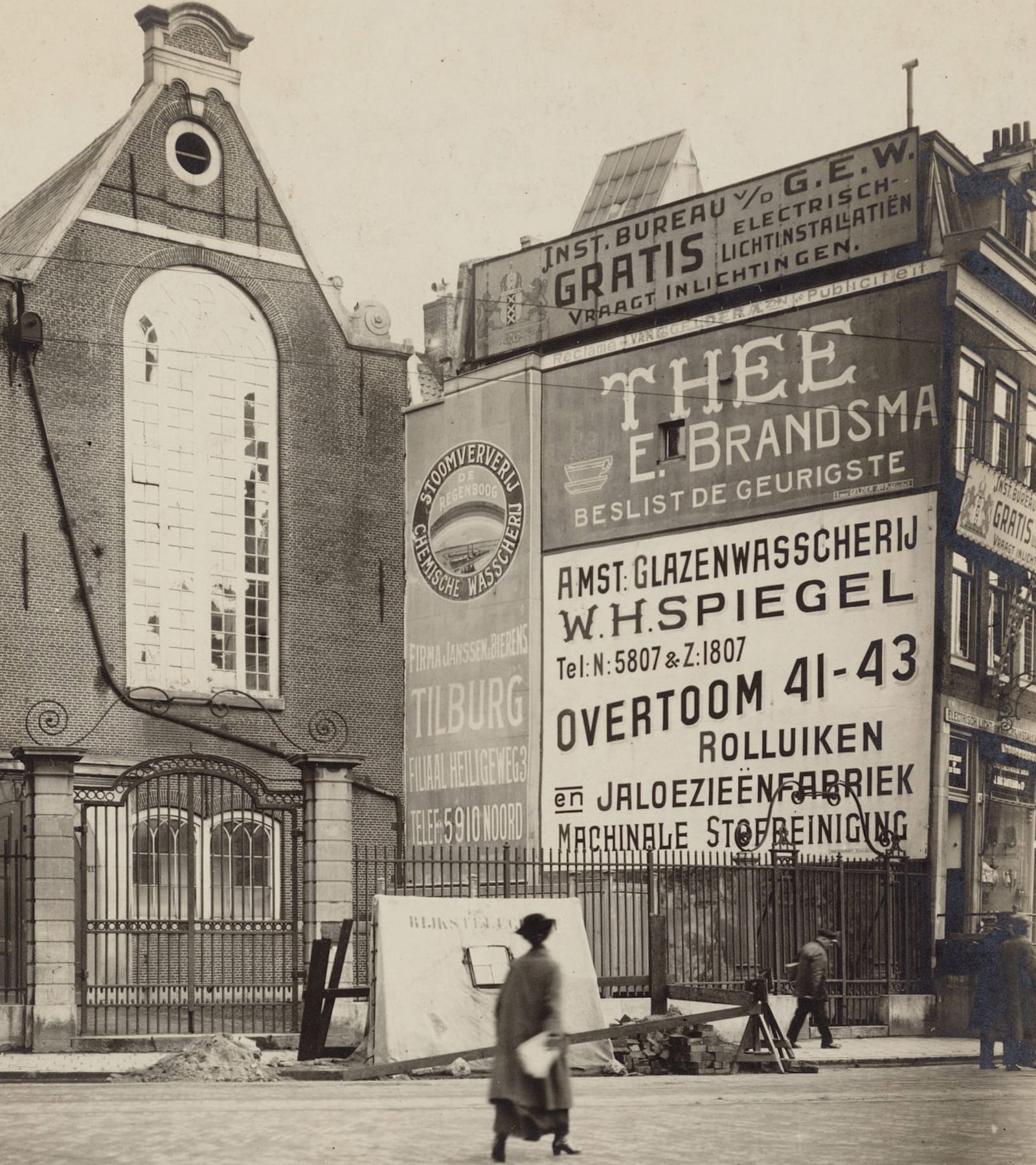 Black and white photo of a wall facing the entranceway to what looks like a church. The wall is almost completely covered by large hand-painted advertisements.