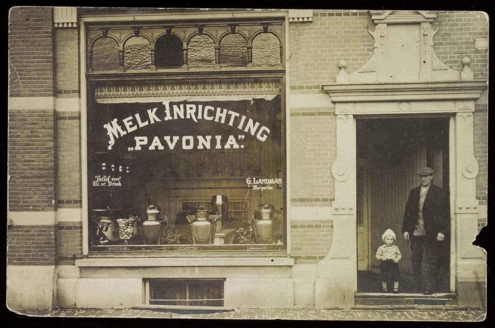 Black and white photo of a building front with a man and toddler standing in the doorway. A large window beside that has a display of urns of various sizes and white lettering on the glass that reads "Melk Inrichting Pavonia".