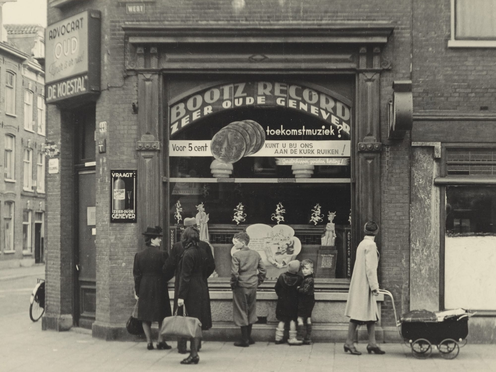 Black and white photo of a corner shop with a few people, including children, gathered outside and apparently discussing the contents of the window display. Signage on the shop advertises "Bootz Record Zeer Oude Genever".