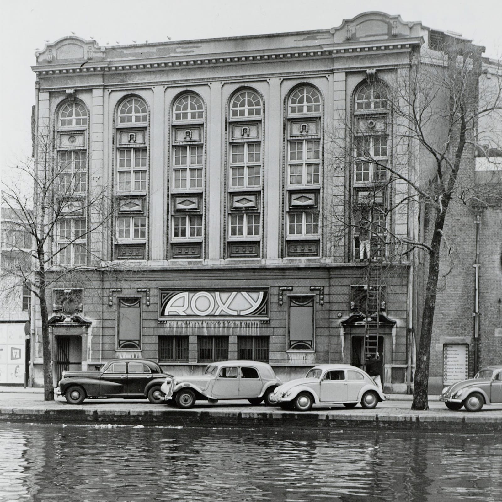 Black and white photo of a grand building viewed from across a canal. A few vintage cars are parked in front of the building that has a large sign that simply says "Roxy" on the frontage.
