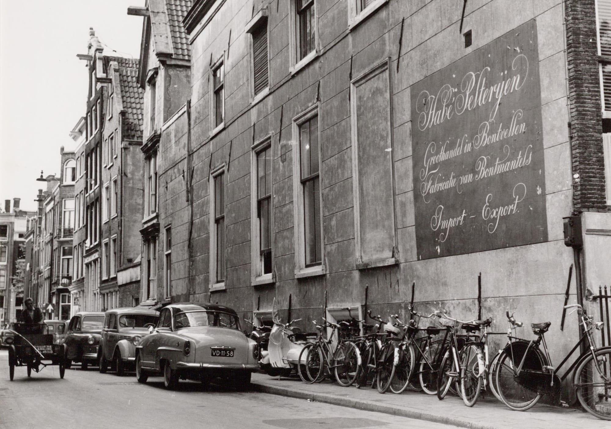 Black and white photo of the view down a street with vintage cars and bicycles parked along it. In the foreground there is a large portion of wall covered with a square advertisement formed of ornate painted cursive letters.