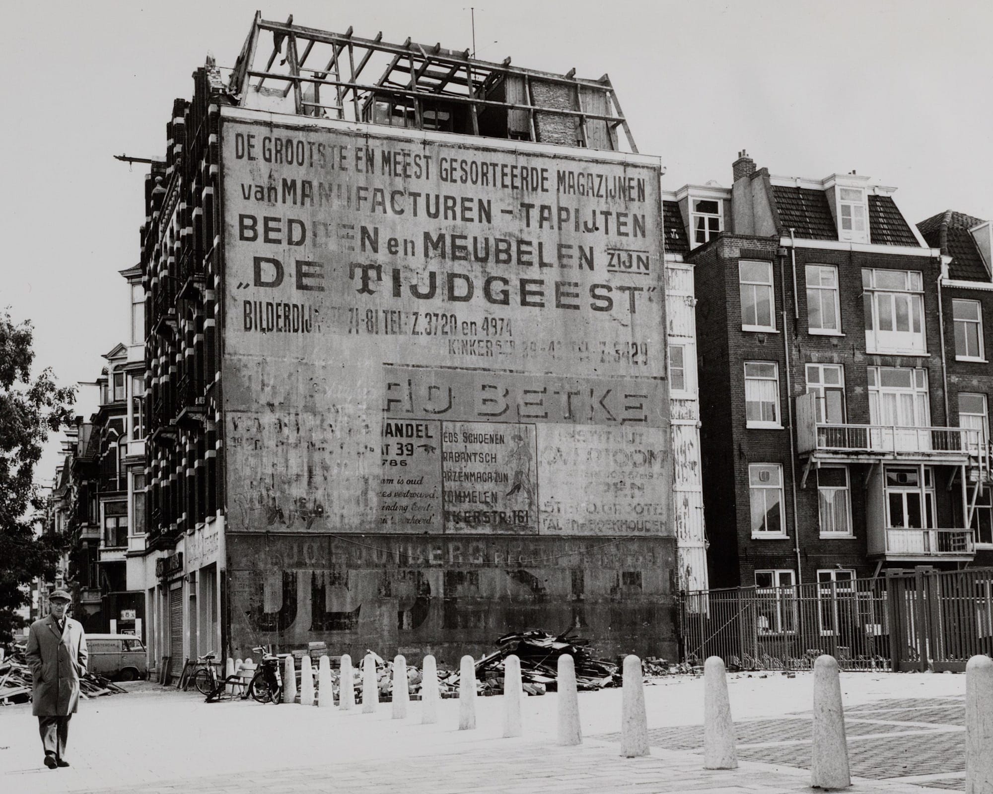 Black and white photo of a derelict building whose facing wall is covered with numerous painted signs that are faded and overlapping with earlier signs.
