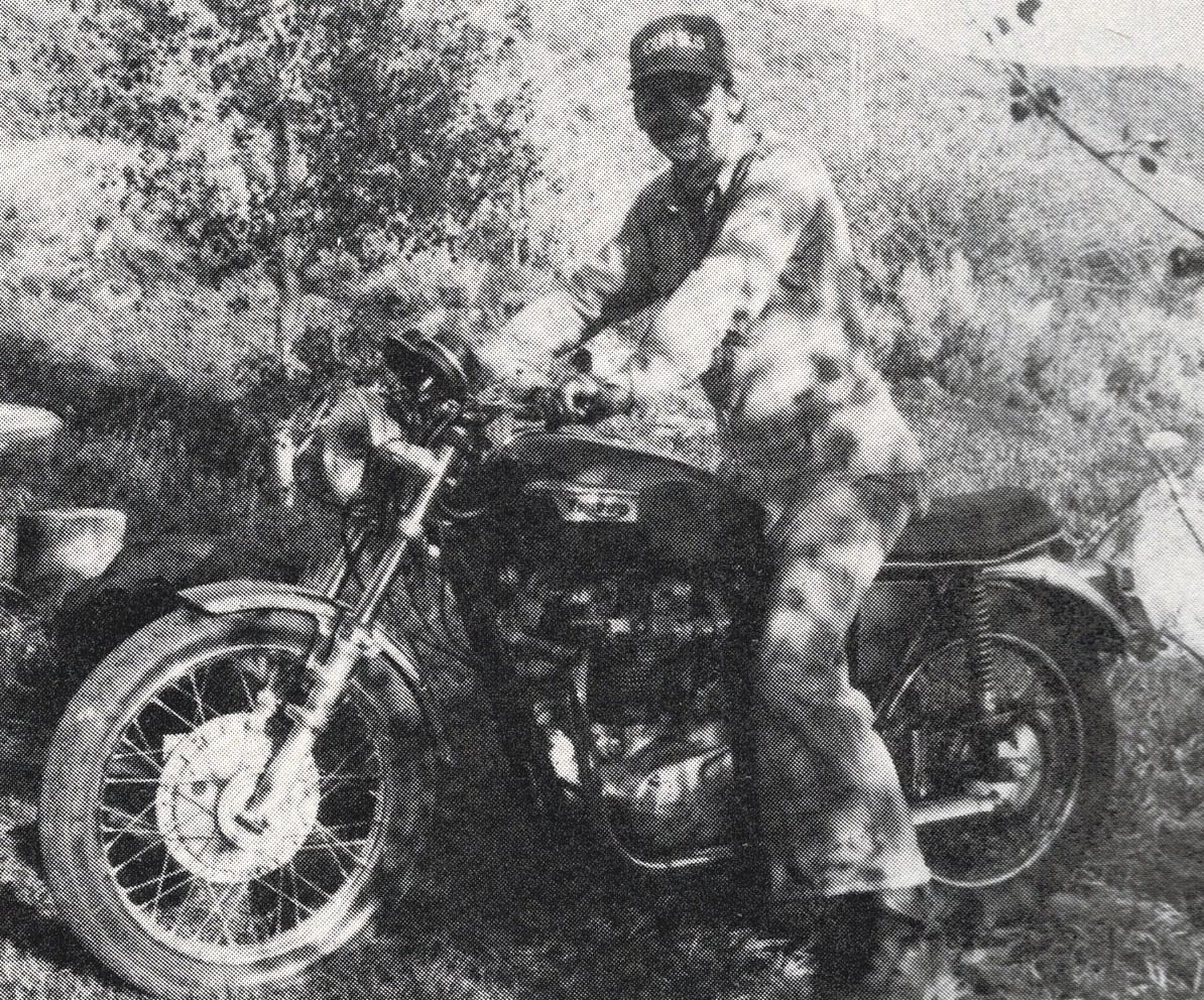 Black and white photo of a man posing on a motorbike dappled in shadows cast by a tree.
