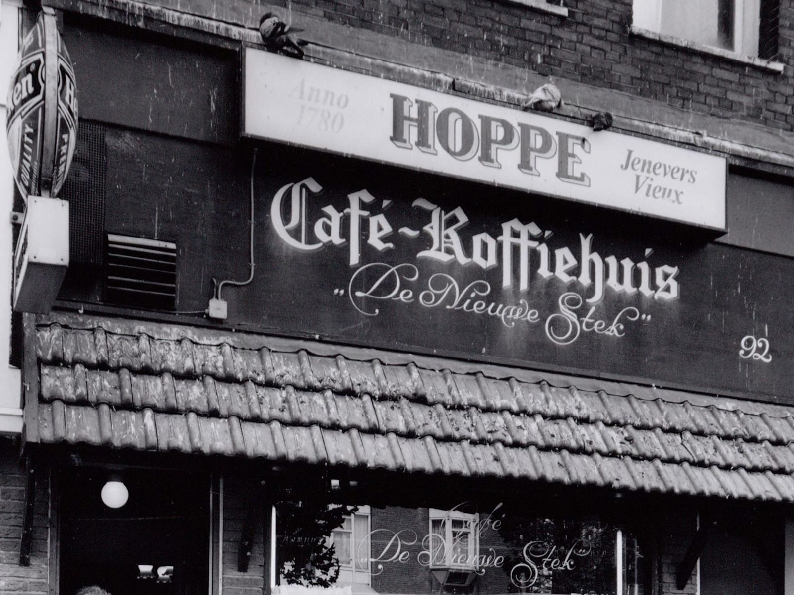 Black and white photo of a shopfront with the focus being the various pieces of signage including the fascia — "Cafe-Koffiehuis, De Nieuwe Stek" — and window. 
