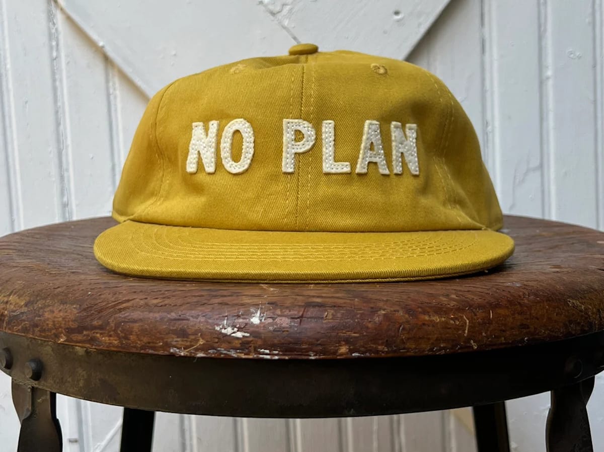 Yellow baseball cap with white stitched-on letters that say "No Plan". It is set on a well-worn wooden stool top.