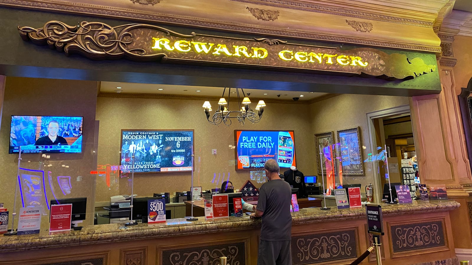 Customer service desk with TV screens behind it. Above is an illuminated sign that says "Reward Center" and framed by the wooden panel with scrollwork at each end, but with the scrolls on the right missing.