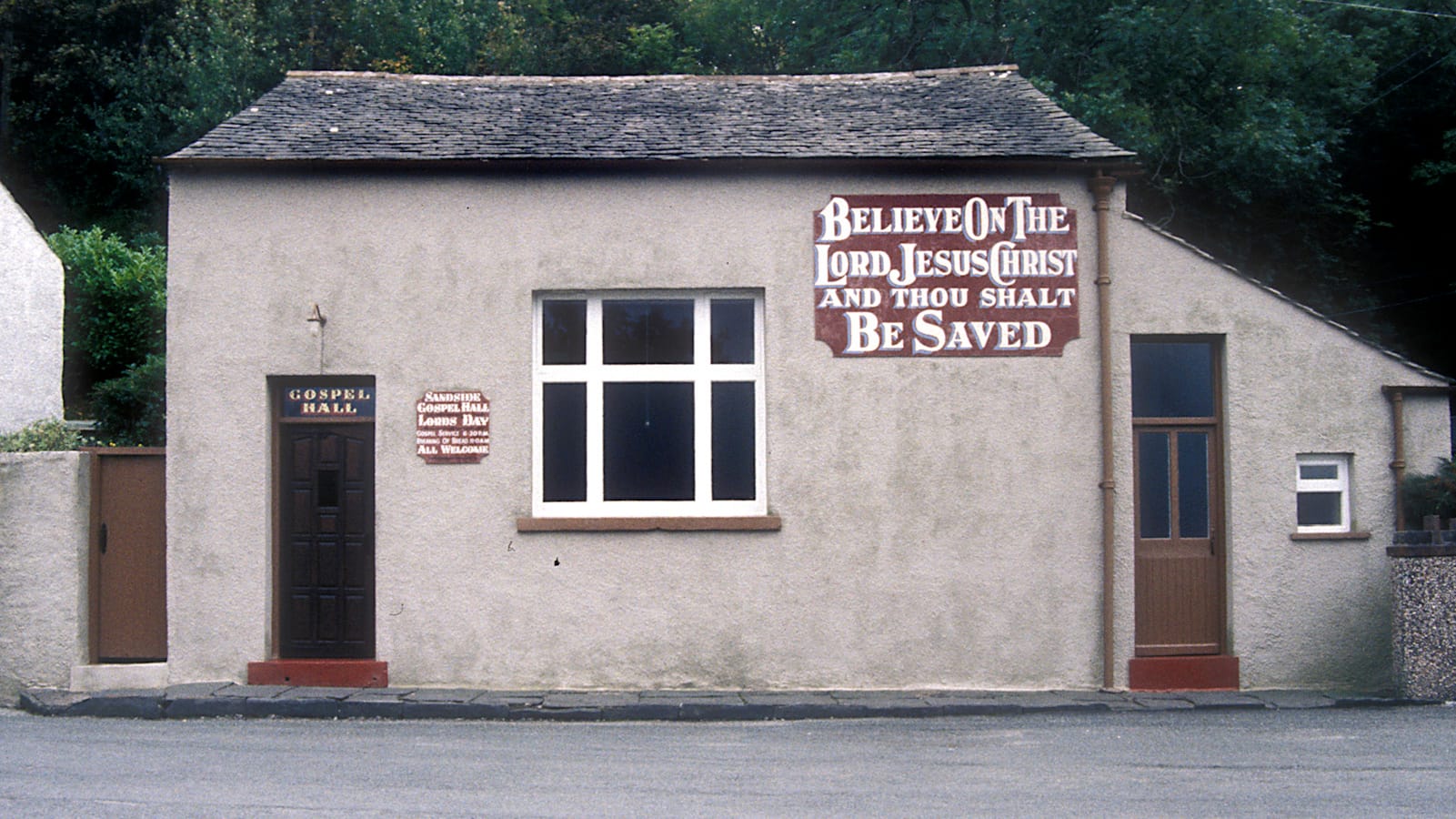 Photo of a simple rural house with a gilded transom that reads "Gospel Hall" above the main door. The plain stucco wall then has two hand-painted panels. The larger of these, one top-right, reads "Believe on the Lord, Jesus Christ, and thou shalt be saved", while the smaller one, between the door and the single window, says "Sandside Gospel Hall" followed by details of services and "All Welcome".