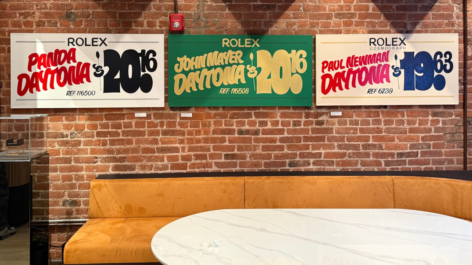 Exposed brick wall with a long tan sofa and marble table in front. Mounted onto the wall are three wide-format painted signs with casual grocery store style lettering. Each one is titled "Rolex" with a model on the left side (Panda Daytona, John Mayer Daytona, and Paul Newman Daytona respectively) and a price on the right, where the price is actually the year of the watch in the grocery store style, e.g. $20.16 for the Panda Daytona.