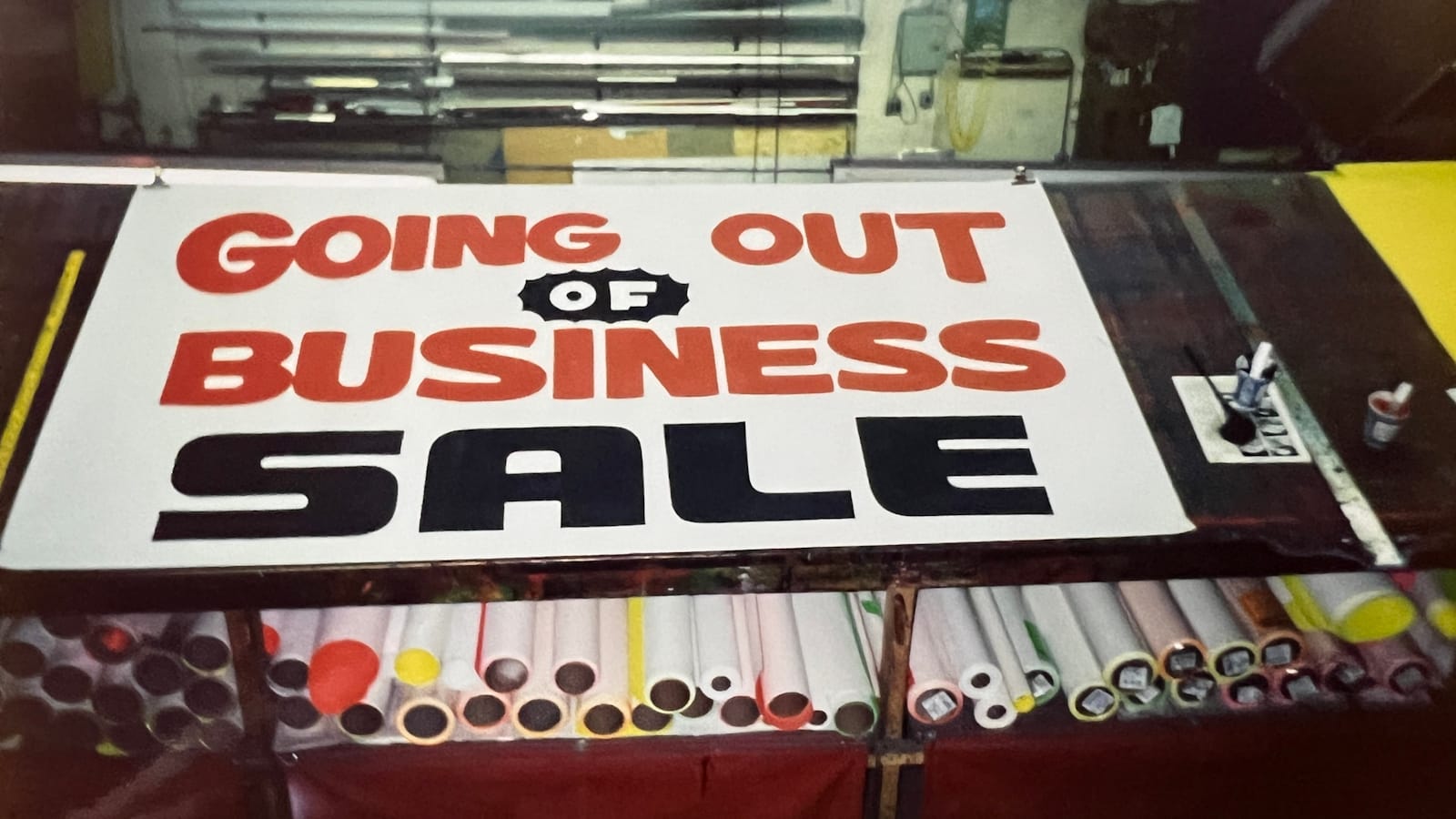 Workbench with lots of rolls of paper stored underneath. On the top is a freshly painted paper sign with simple lettering in red and black that reads "Going out of business sale".
