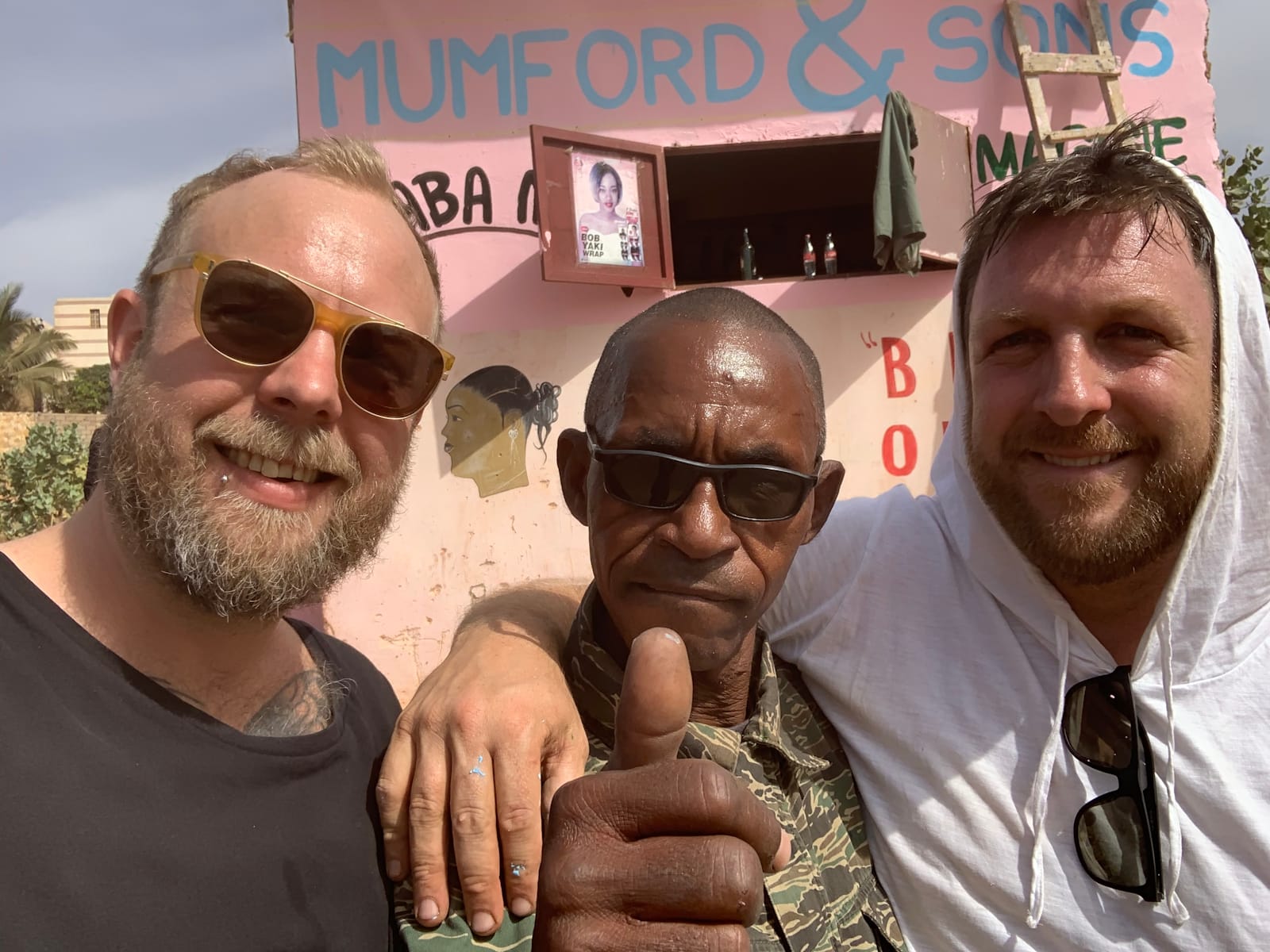 Three men posing for a photo. The one in the middle is giving a thumbs up and wearing a neutral expression, while the two either side are smiling, with the one on the right having his arm over the shoulder of the one in the middle. Behind them is a small simple building with a pink wash and the lettering on it for the EP title and artist names.