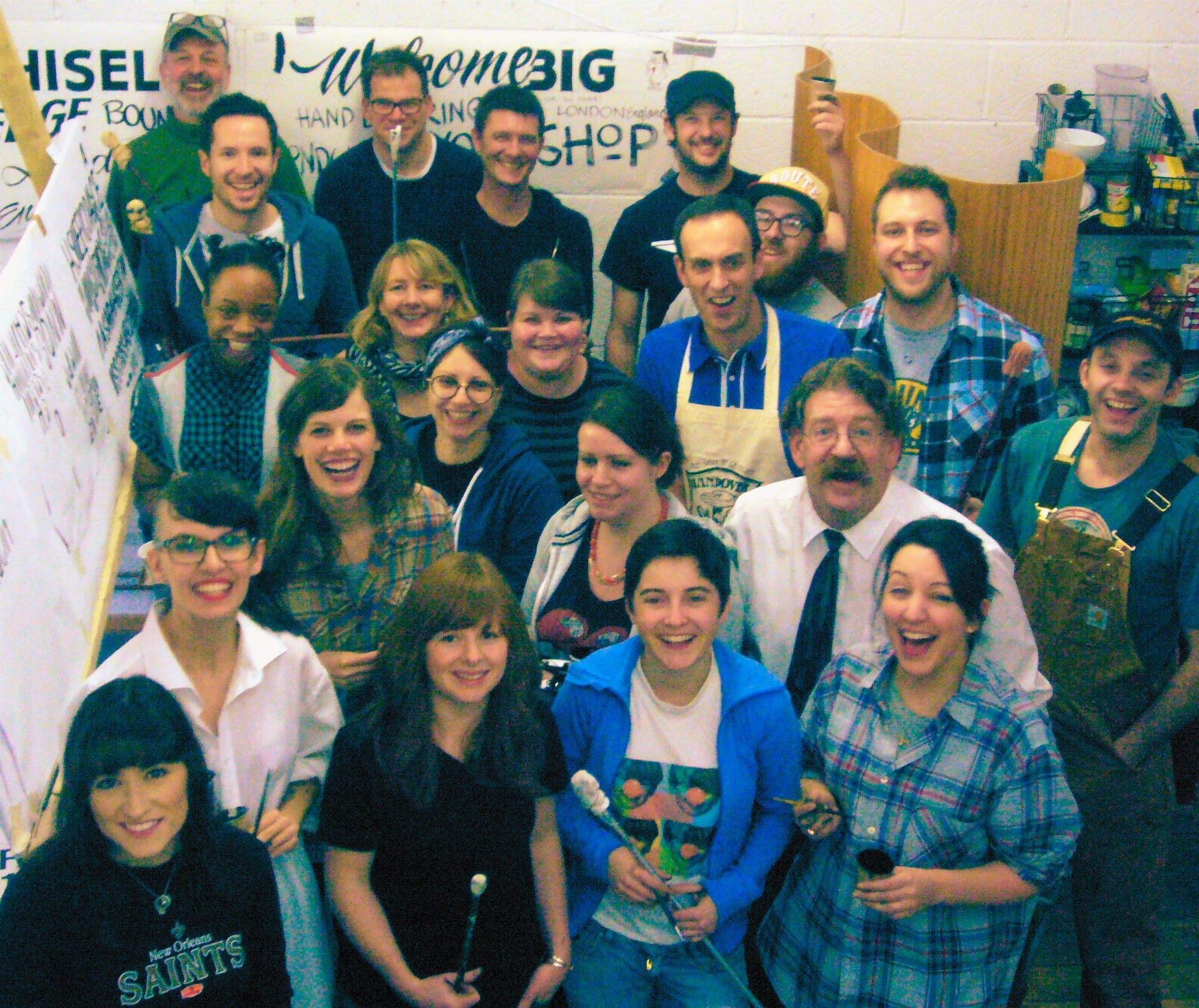 Group photo taken from above of a smiling crowd of about 20 people, some of whom are holding tools and materials used in sign painting.