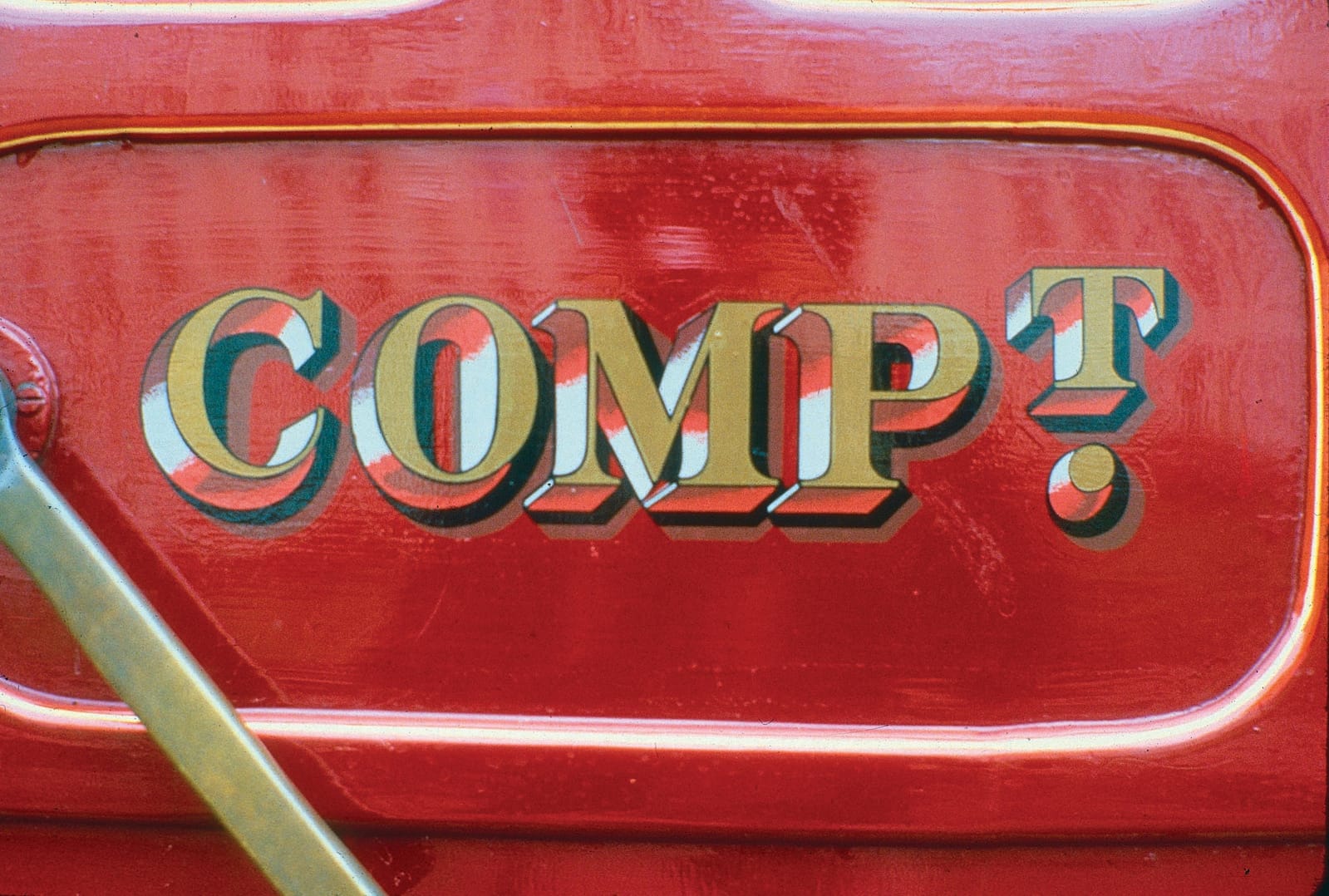 Bright red part of a vehicle with the lettering in gold, blocked with Red and white, with black and brown cast shadow. It says "Comp't", which would appear to be an abbreviation of compartment.