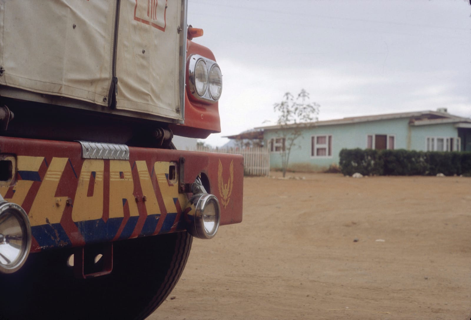 Dusty road scene with a building in the background and a portion of the back of a truck in the foreground. A low-down panel on the back of the truck has the rear lights set within in it and, between those, lettering that says "Gloria".