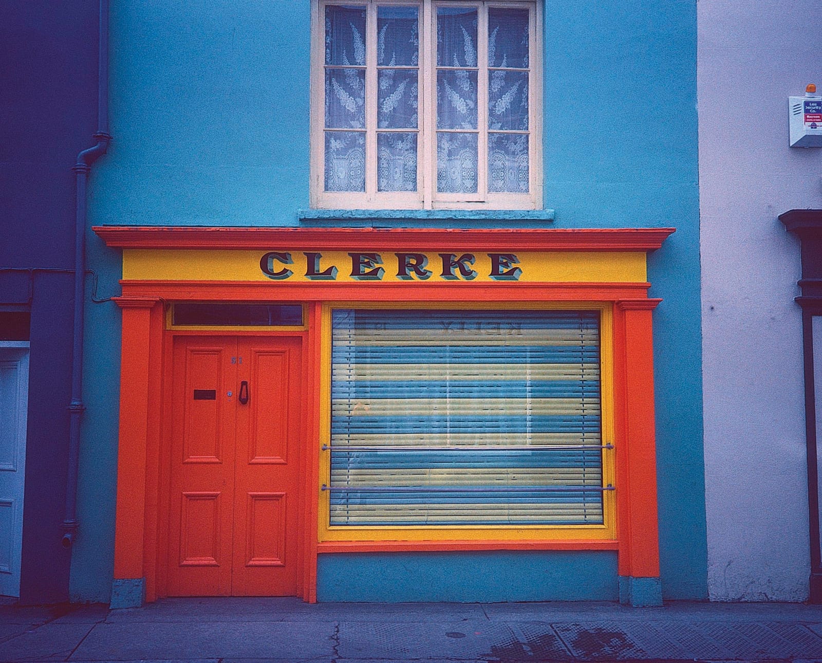 Simple shop front on a terrace with the building painted blue and the fittings plus door and window in brighter yellow and orange. Lettering in dark blue with a light blue shade fill the middle part of the fascia board and reads "Clerke".
