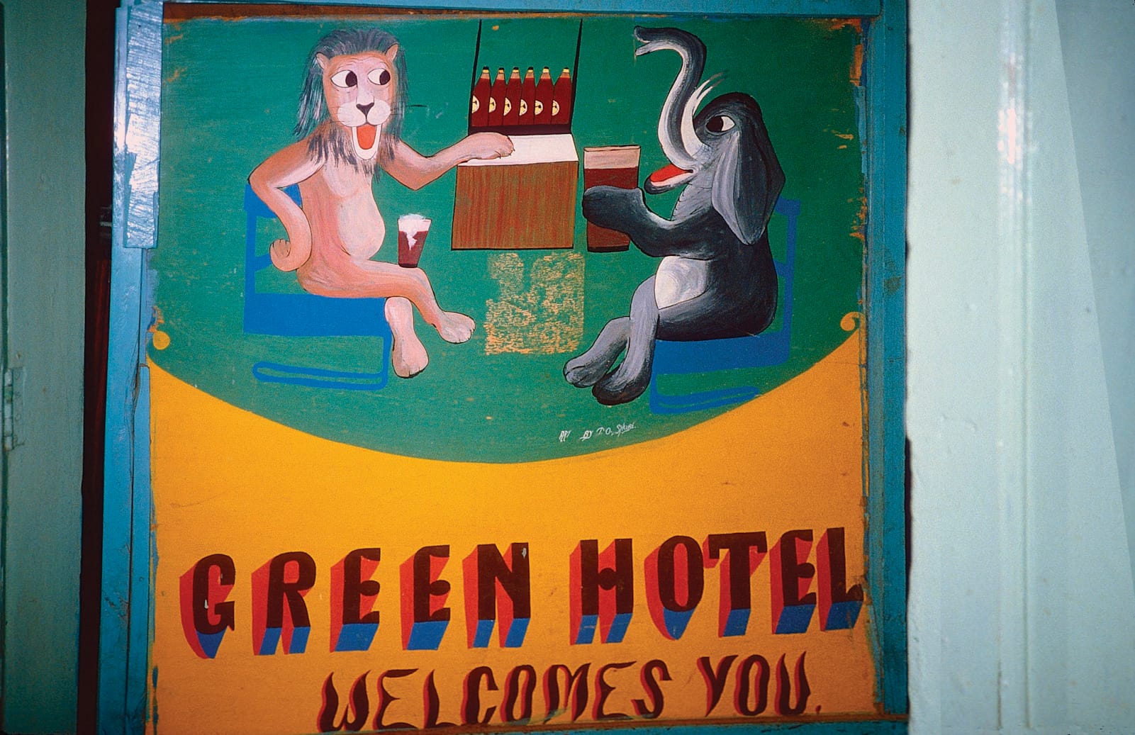 Painted mural sign with a pictorial showing a lion and an elephant seated at a bar drinking beer. Lettering below this says "Green Hotel Welcomes You".