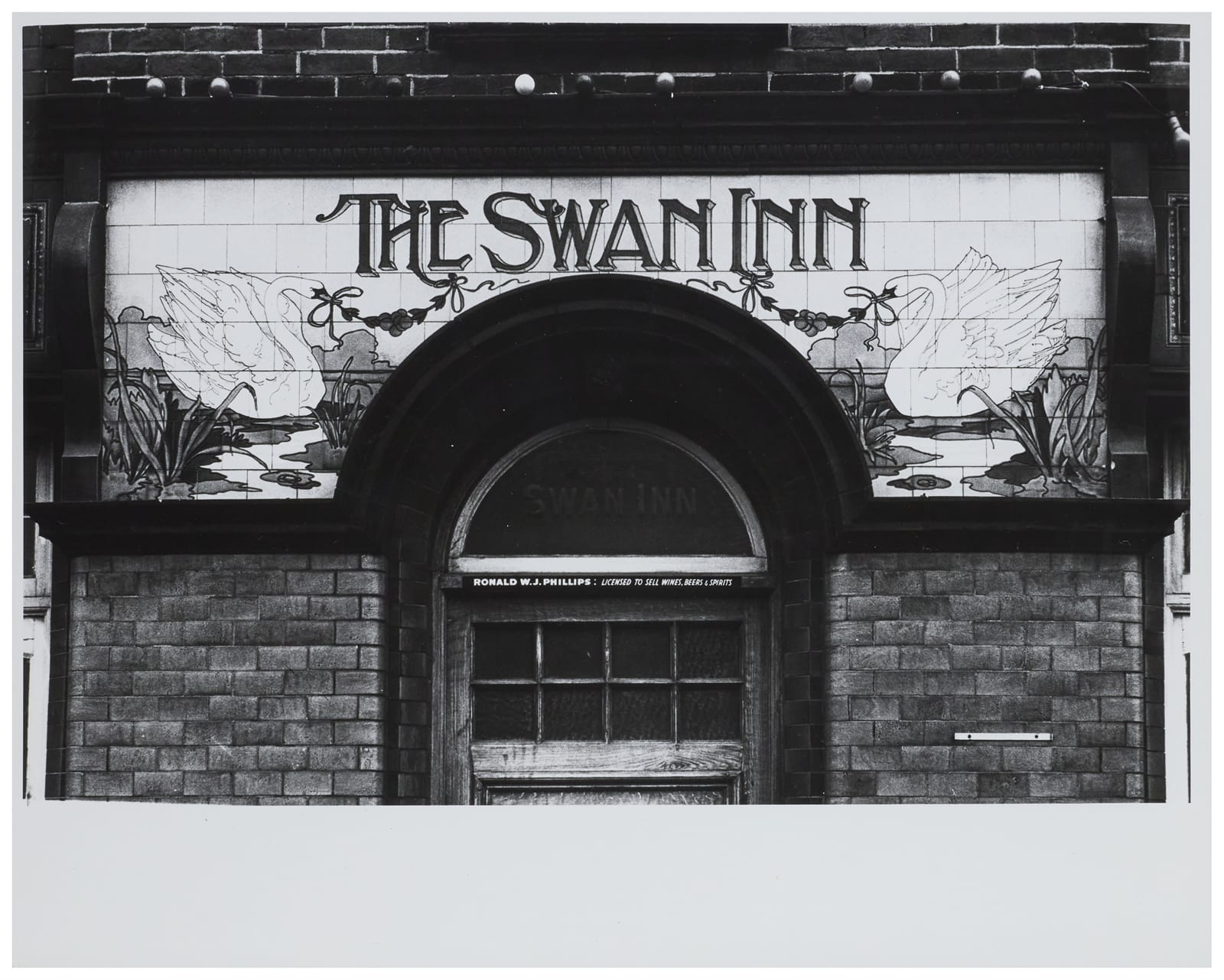 Ceramic tile sign set around a semi-circular tramsom window. The upright decorative lettering for "The Swan Inn" is flanked by pictorials of swans left and right.