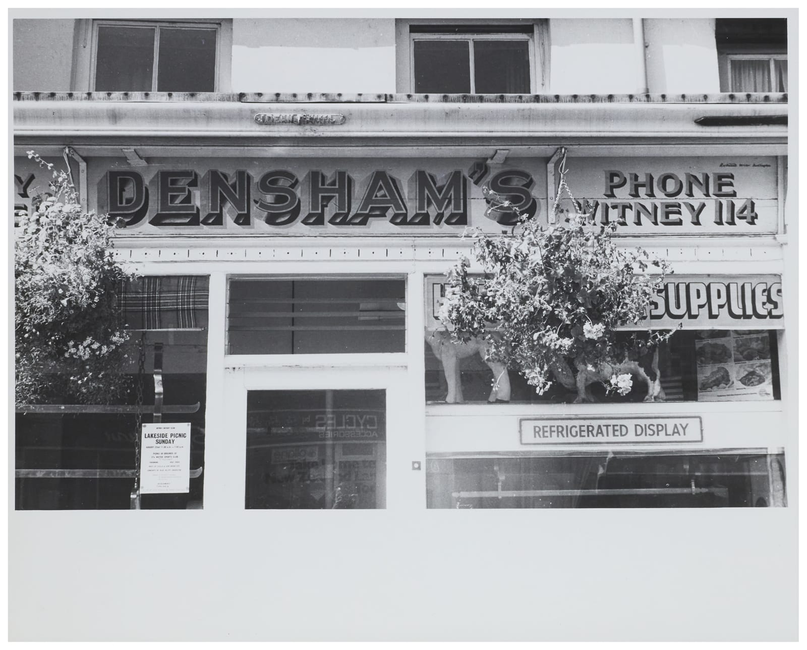 Black and white photo of a shopfront with hanging baskets and hand-painted dimensional lettering that says "Densham's" and a smaller "Phone Witney 114". 
