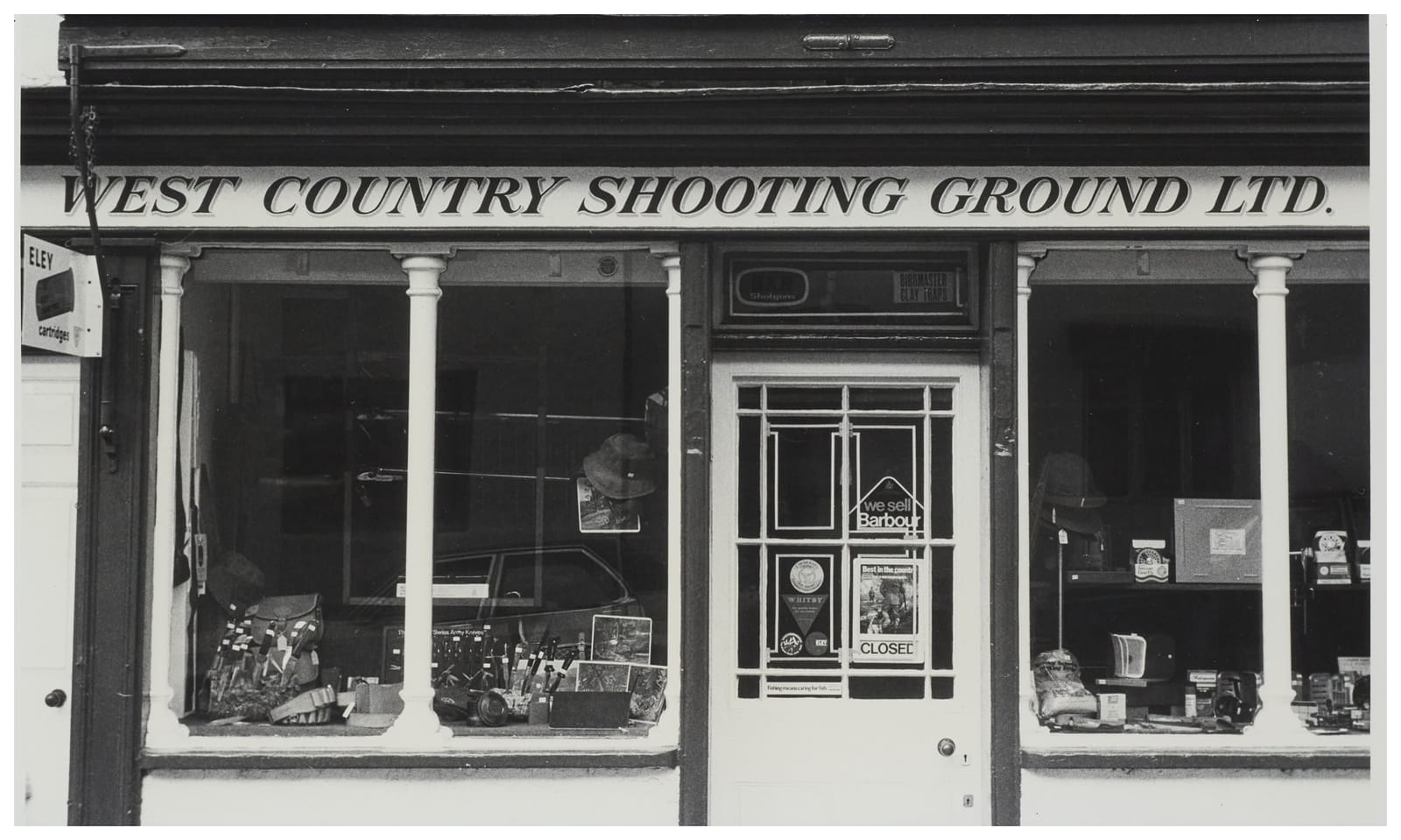 Shopfront with window displays. The fascia sign has simple italic serifed capitals in black with a simple drop shade saying "West Country Shooting Groud Ltd."