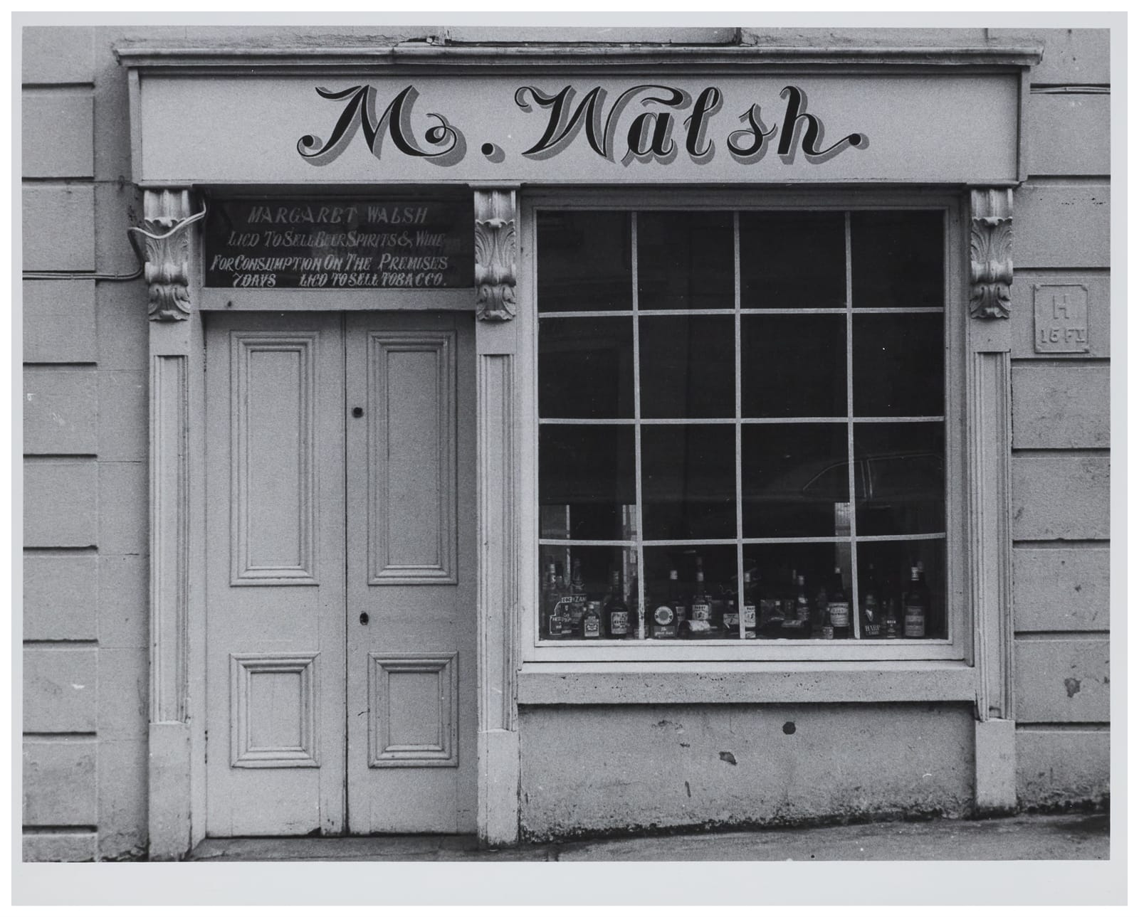 Photo of a simple shopfront with bottles in the window. The fascia sign says "M. Walsh" in italic lettering that doesn't join and has varying spacing between letters. The transom window has four lines of lettering giving the licensee details. 