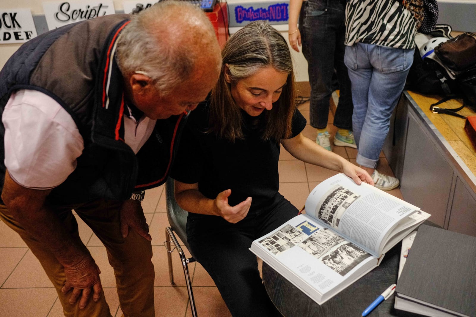 Older man bending over to look at a spread from a book that a seated woman has open as she gestures while speaking with a smile on her face.
