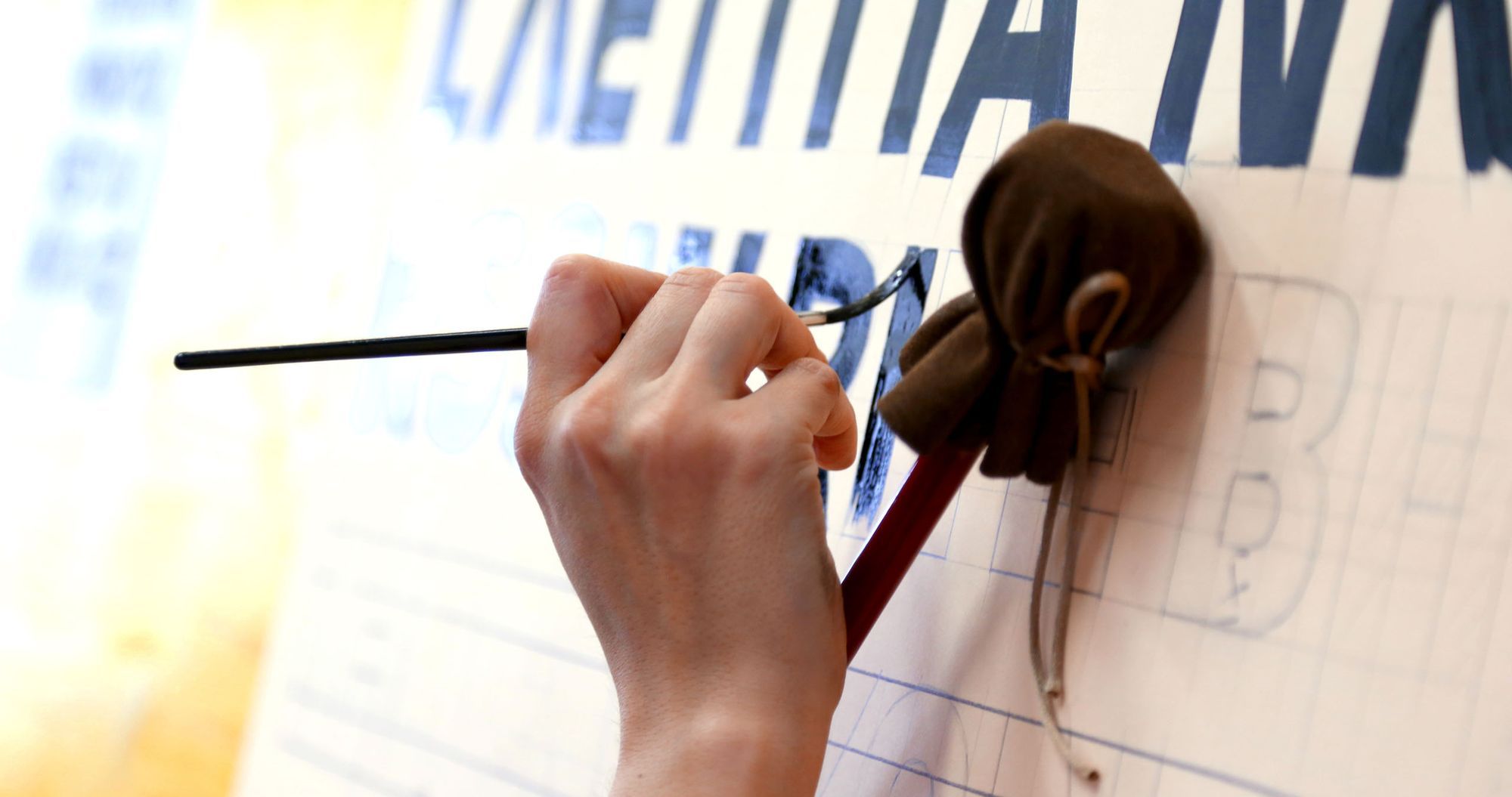 Hand resting on a mahl stick while holding a brush and painting letters in black.