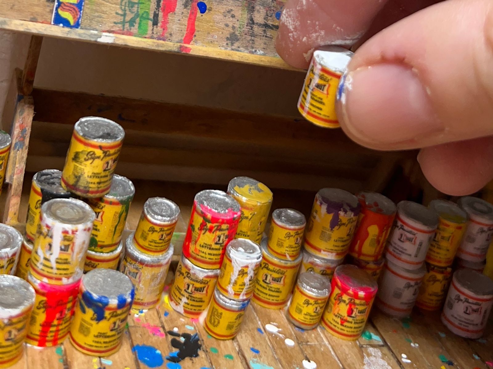 Close-up of a hand picking up a tiny tin of paint made to scale for a larger diorama.