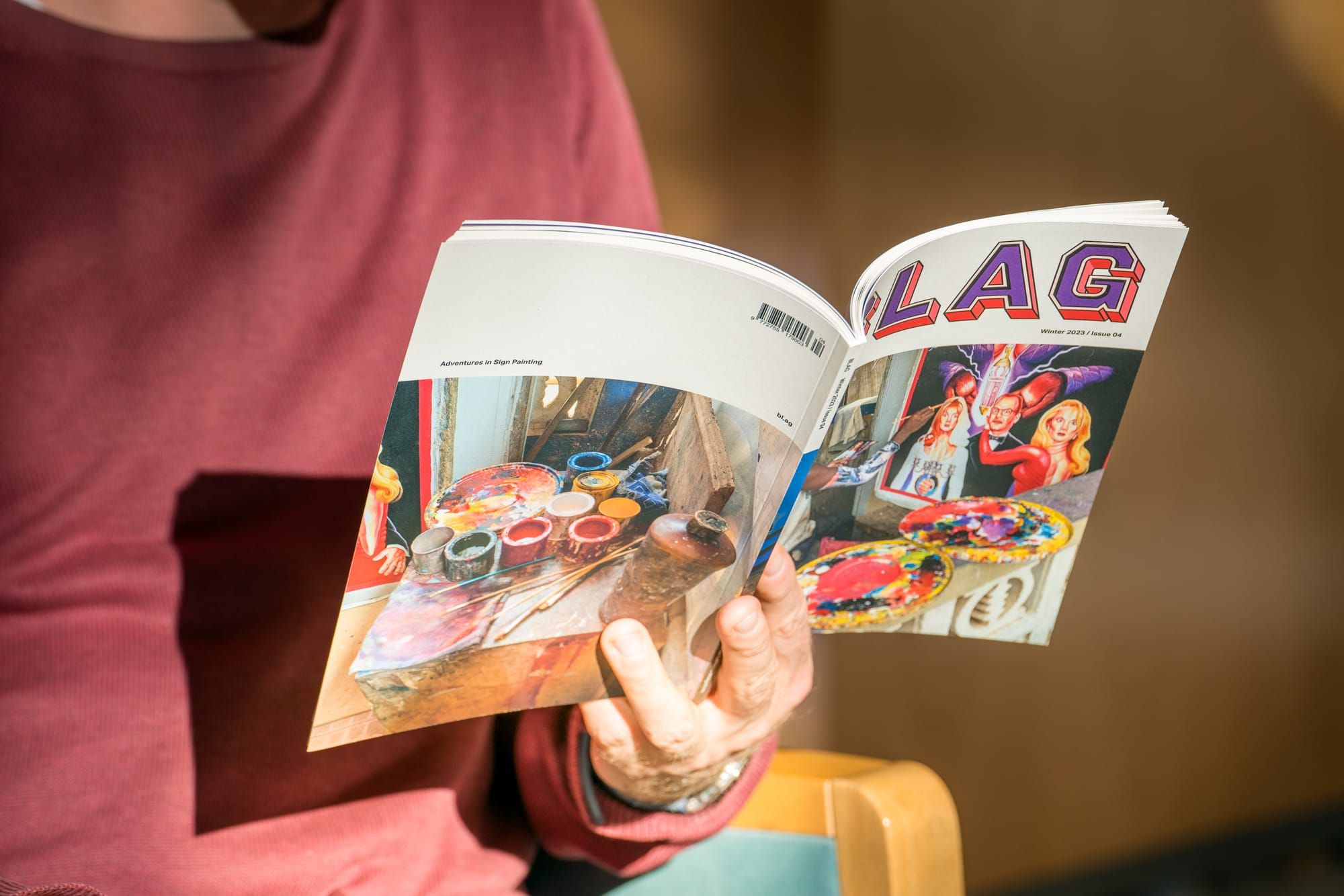 Hand holding a magazine with the person's torso visible behind in a burgundy jumper.