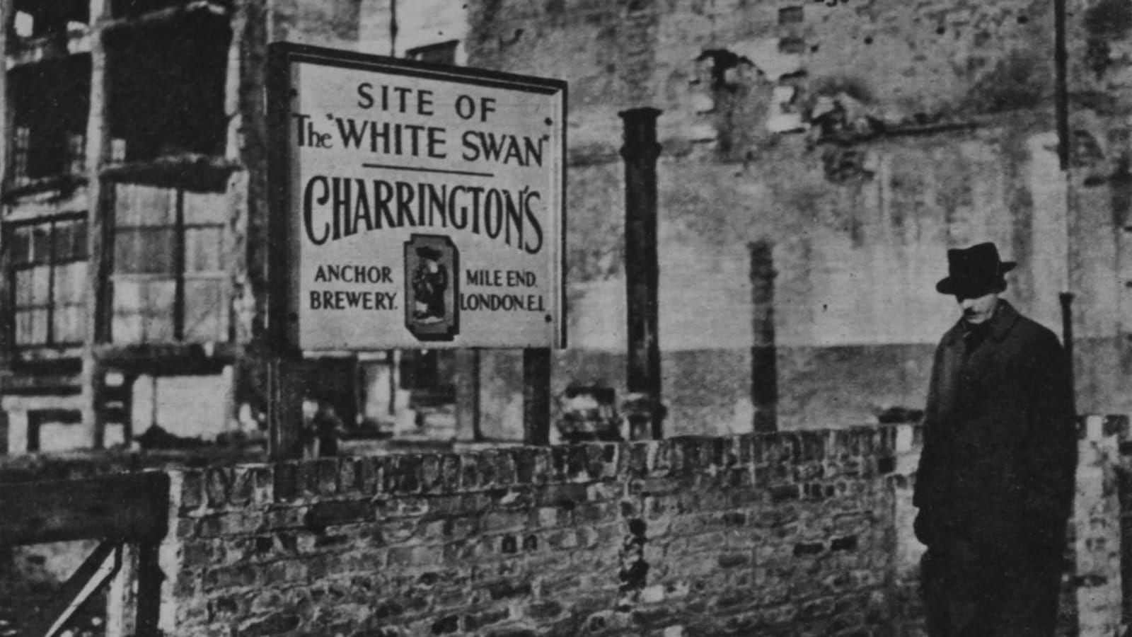 Man in a hat in front of a wall and a hand-painted sign with destroyed buildings behind.