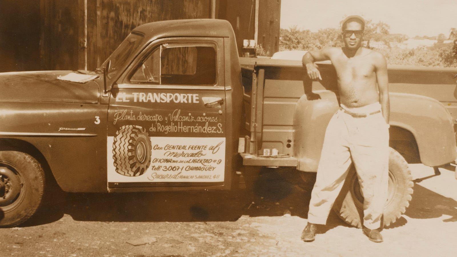 Sepia photo of a topless man resting his arm on the back of a truck that has hand-painted lettering on the door.