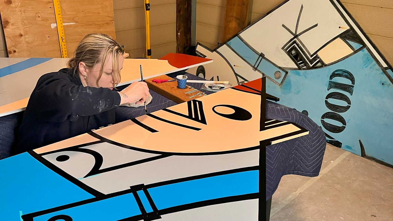 Woman sandwiched between two sheets of painted plywood, and painting Mr. Plywood's face onto one of these. 