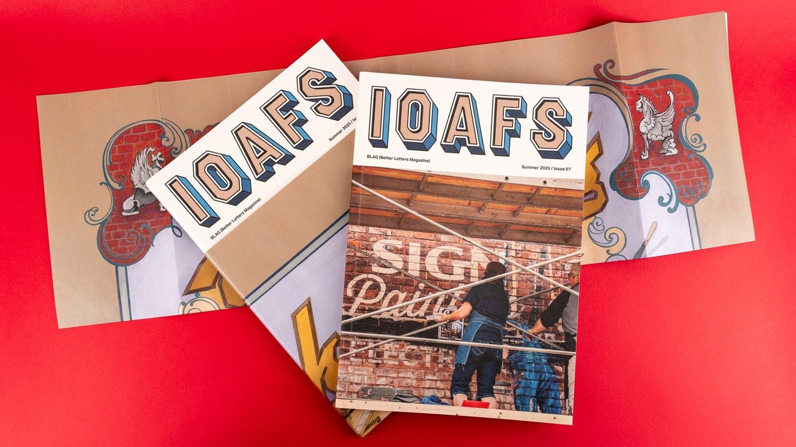 Two magazines with masthead IOAFS placed on top of a folded poster which has been removed to reveal the cover on one copy of the magazine.