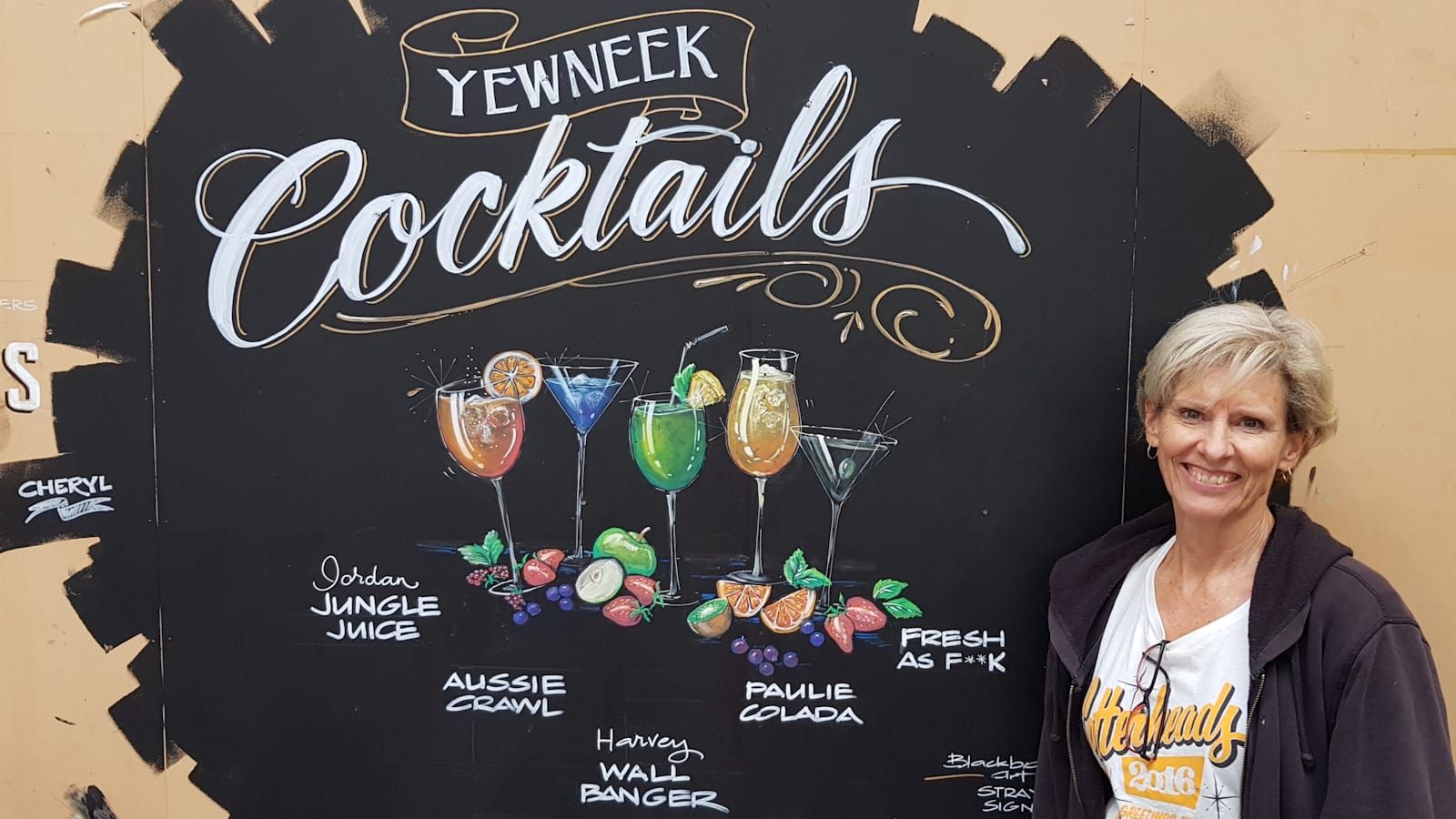 Woman standing in front of a hand-drawn and lettered blackboard advertising a variety of colourful cocktails.