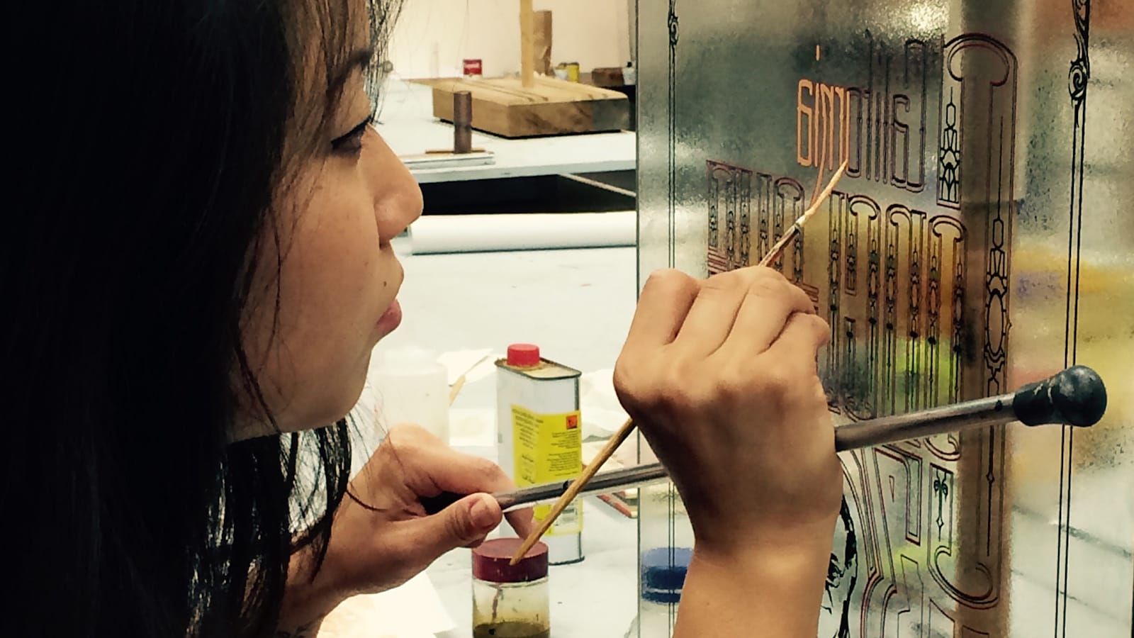 View from the side of a woman working with a mahl stick and fine brush to paint lettering on the back of a piece of glass.