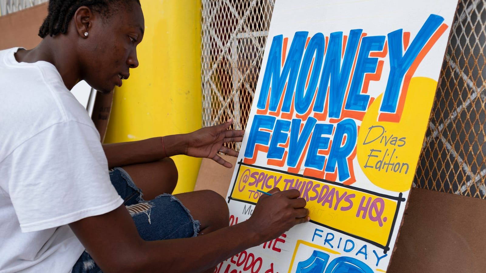 Man sat on a stool painting a sign advertising "Money Fever, Divas Edition, at Spicy Thursdays HQ."