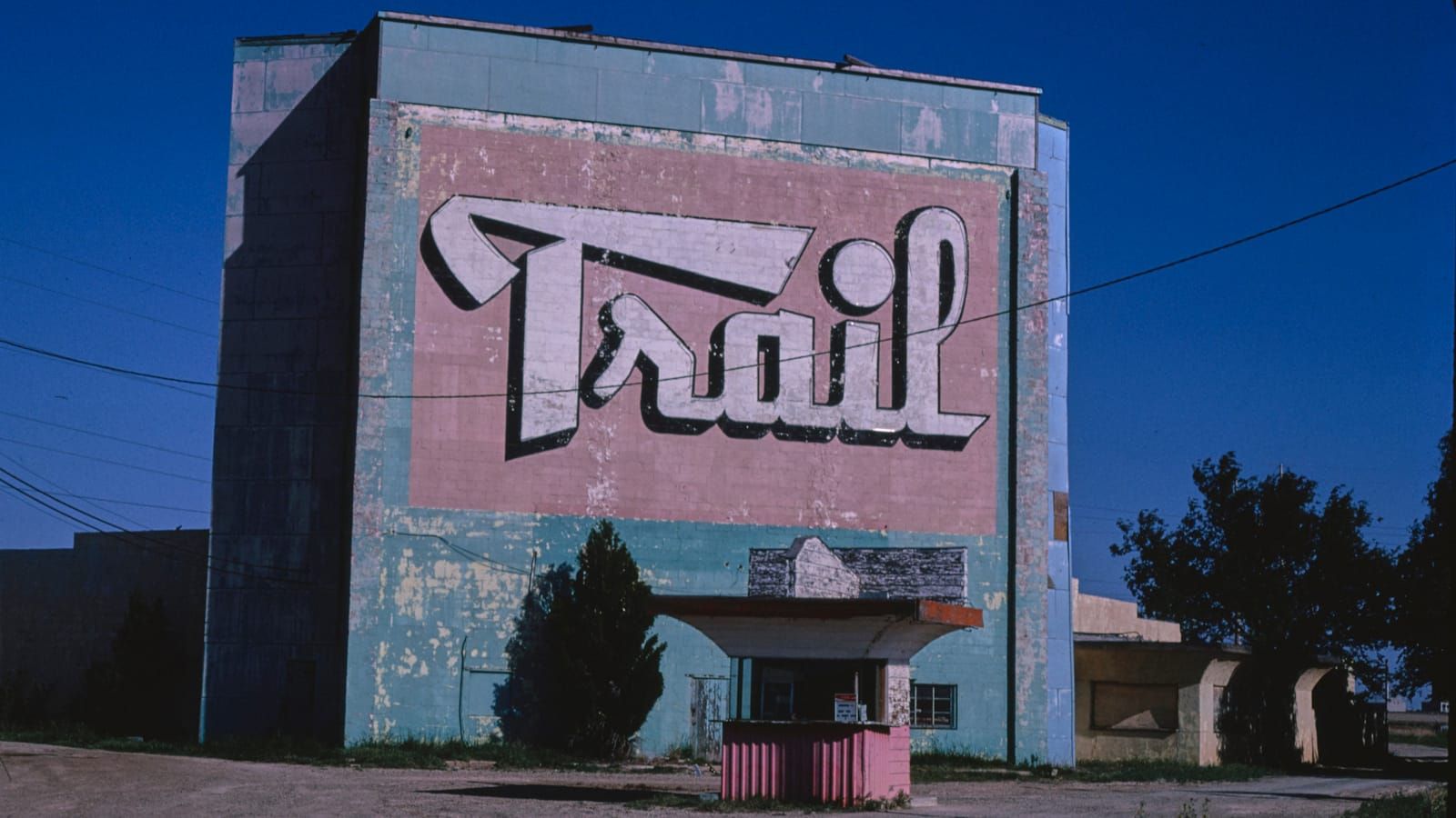 Large building with fading and damaged paintwork. A huge hand-painted sign simply reads "Trail" while a ticket kiosk occupies the ground in front.