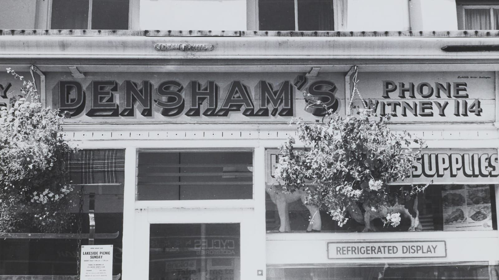 Black and white photo of a shopfront with hanging baskets and hand-painted dimensional lettering that says "Densham's" and a smaller "Phone Witney 114". 