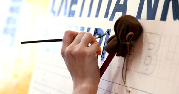 Hand resting on a mahl stick while holding a brush and painting letters in black.