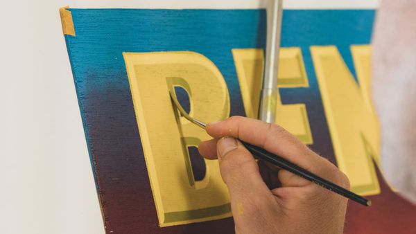 Close-up of a sign painter adding a shade to a hand-painted letter 'B' in yellow paint.